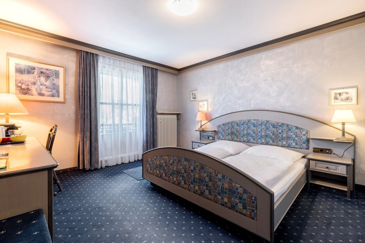 Une chambre d’hôtel comprenant un lit, des tables de chevet, un bureau, une chaise et une fenêtre avec des rideaux.