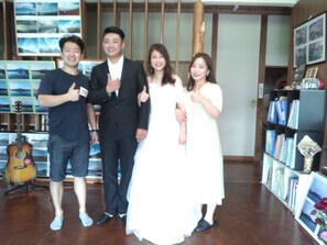 A group of people posing in a room with a bookshelf and a guitar. - Ryokan Fuji Heights (Fujikawaguchiko)