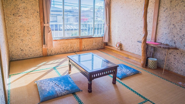 A tatami mat room with a glass table and cushions, a window with a view, and a small shelf with a picture. - Ryokan Fuji Heights (Fujikawaguchiko)