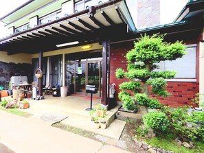 A building with a covered entrance, a brick wall, and a well-maintained garden with potted plants and a bonsai tree. - Ryokan Fuji Heights (Fujikawaguchiko)