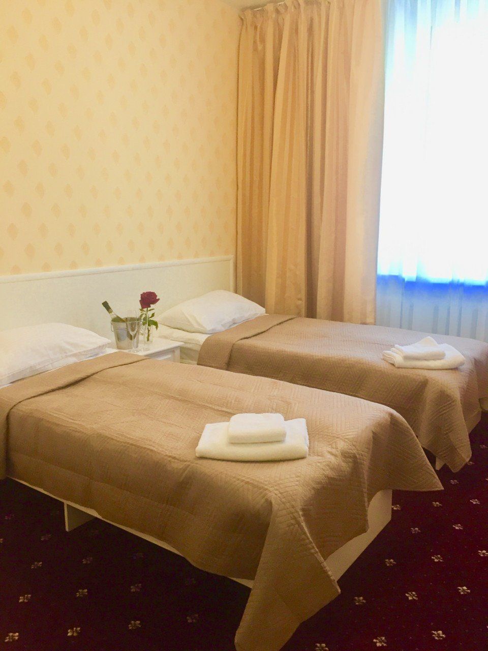 A hotel room with two beds, each with a white pillow and a folded towel. There is a small table with a vase containing a single rose. The room has a patterned wallpaper and a window with curtains.