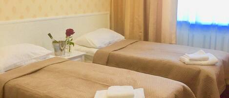 A hotel room with two beds, each with a white pillow and a folded towel. There is a small table with a vase containing a single rose. The room has a patterned wallpaper and a window with curtains.