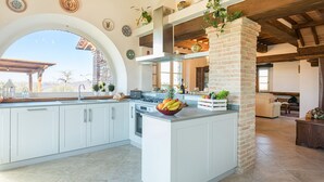 Une cuisine spacieuse avec des meubles blancs, un grand îlot central orné de fruits et une vue dégagée sur l’extérieur grâce à une fenêtre en arc de cercle.