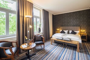 Habitación de hotel con una cama grande, una silla de madera, una mesita con una botella y vasos, y vistas a árboles por la ventana.