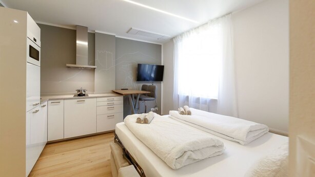 Een moderne hotelkamer met een kitchenette, een flatscreen televisie, een bed met netjes opgevouwen handdoeken en een groot raam met transparante gordijnen.