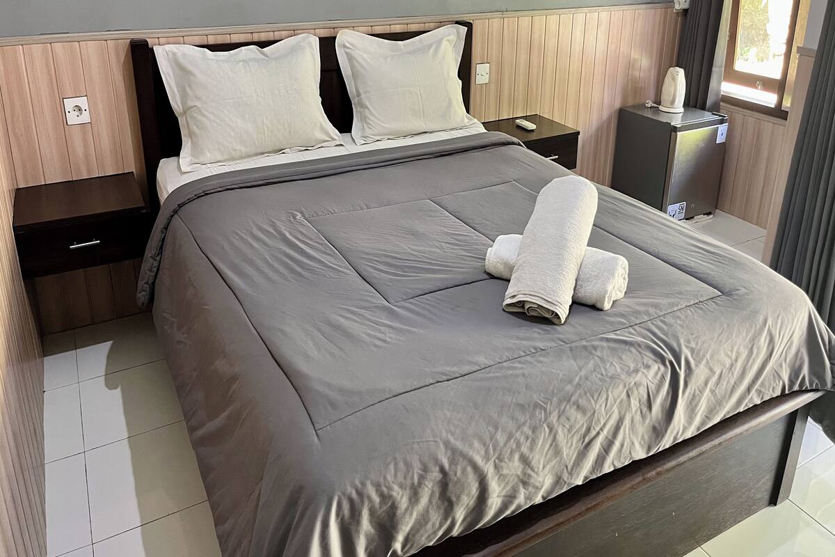 Een netjes opgemaakt bed met een grijze sprei en twee witte kussens, een nachtkastje met een afstandsbediening en een klein kastje met een waterkoker.