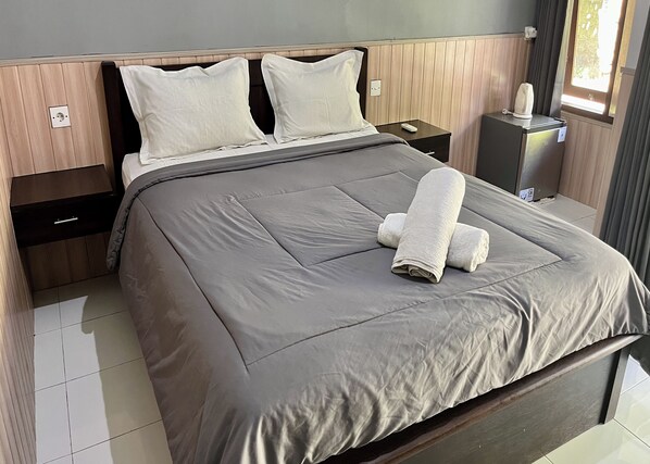 A neatly made bed with a grey bedspread and two white pillows, a bedside table with a remote control, and a small cabinet with a kettle. - Mai Guesthouse Sanur (Denpasar)