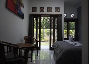 A bedroom with a bed, a chair, and a table. There is a sliding door leading to an outdoor area with greenery. - Mai Guesthouse Sanur (Denpasar)