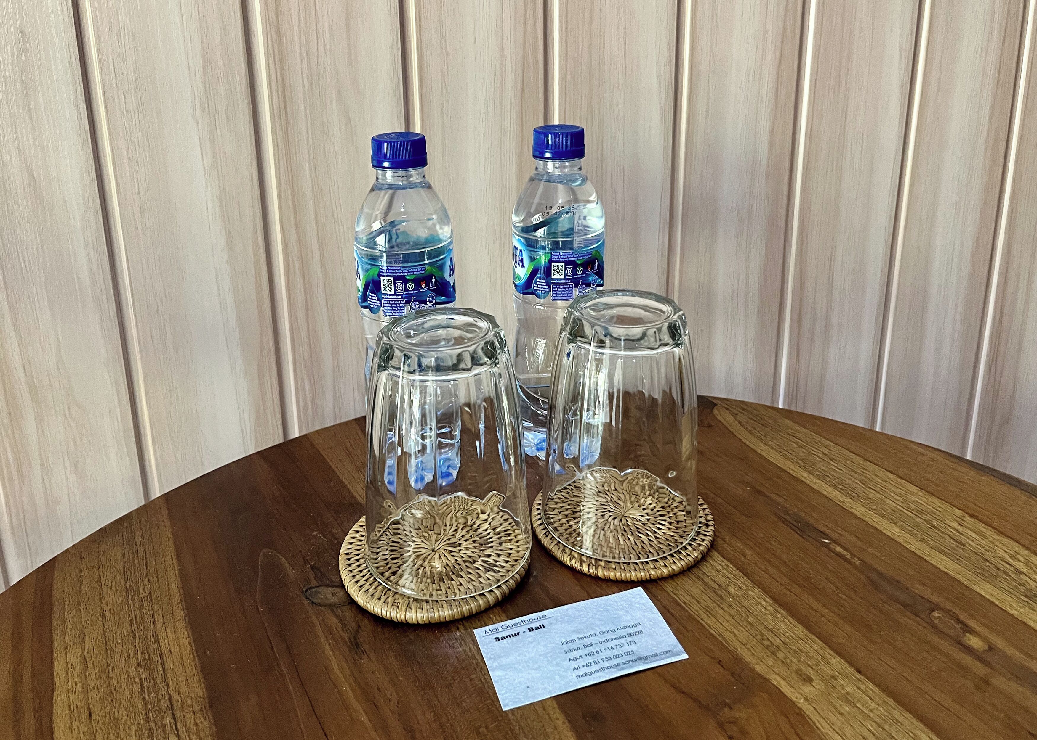 Two water bottles and two clear glass tumblers on a wooden table with a woven coaster, a business card, and a wooden wall in the background.