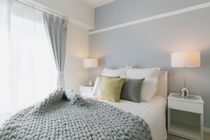 Een moderne slaapkamer met een groot bed, twee nachtkastjes met lampen, en een raam met dunne gordijnen.