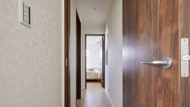 Um corredor com uma porta de madeira, uma cama ao fundo e um interruptor de luz na parede.