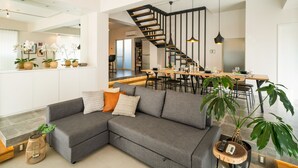 A modern living room with a gray sectional sofa, a wooden coffee table, and a large potted plant. A staircase leads to a dining area with a wooden table and chairs. - Infinity Hotel -Higawa- (Naha)