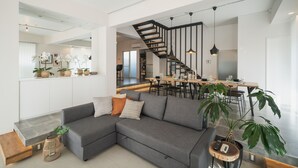A modern living room with a grey sectional sofa, a staircase, and a dining area with wooden tables and chairs. - Infinity Hotel -Higawa- (Naha)