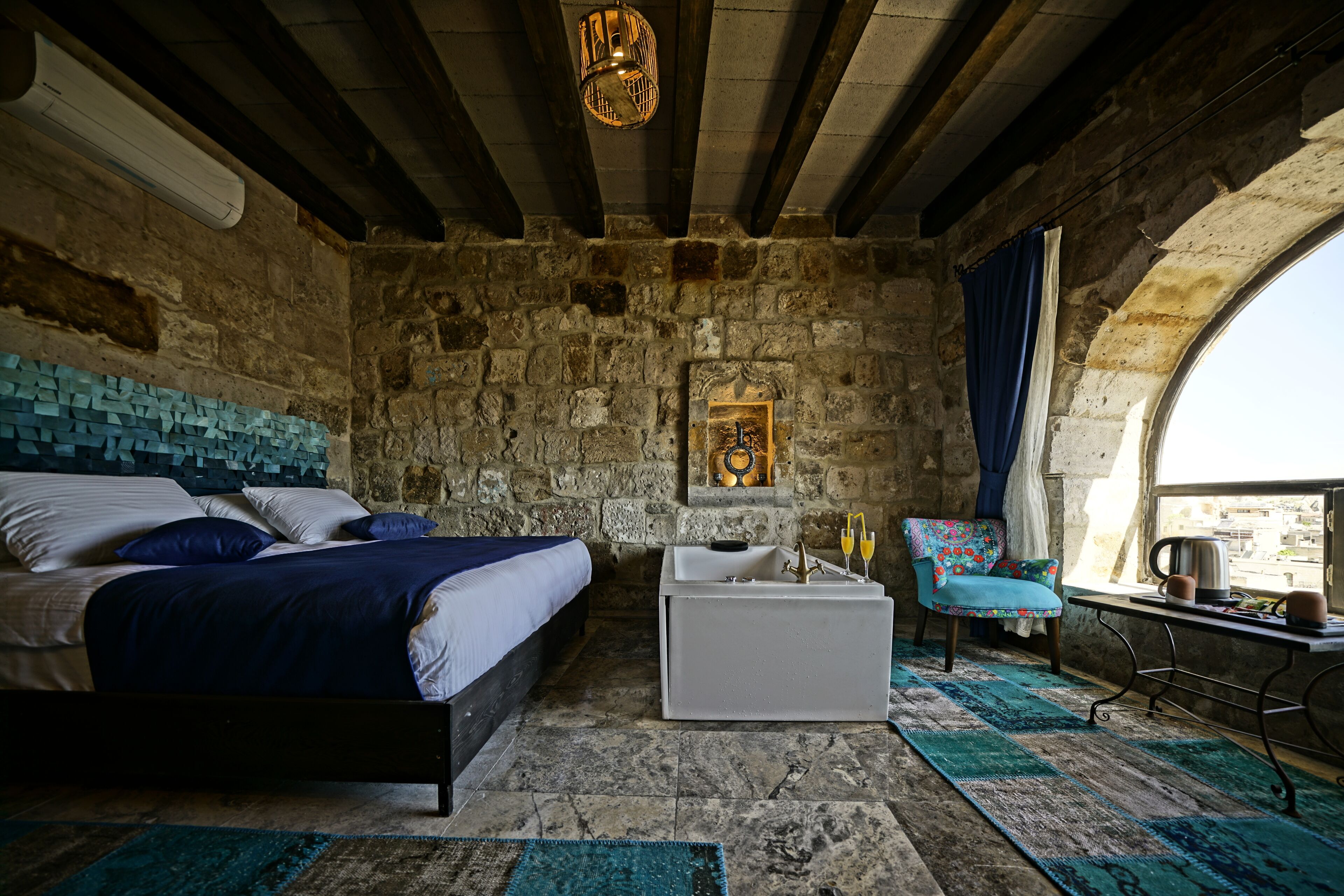 Un dormitorio rústico con pared de piedra, cama con ropa de cama azul, mesita de noche blanca y ventana con vistas.
