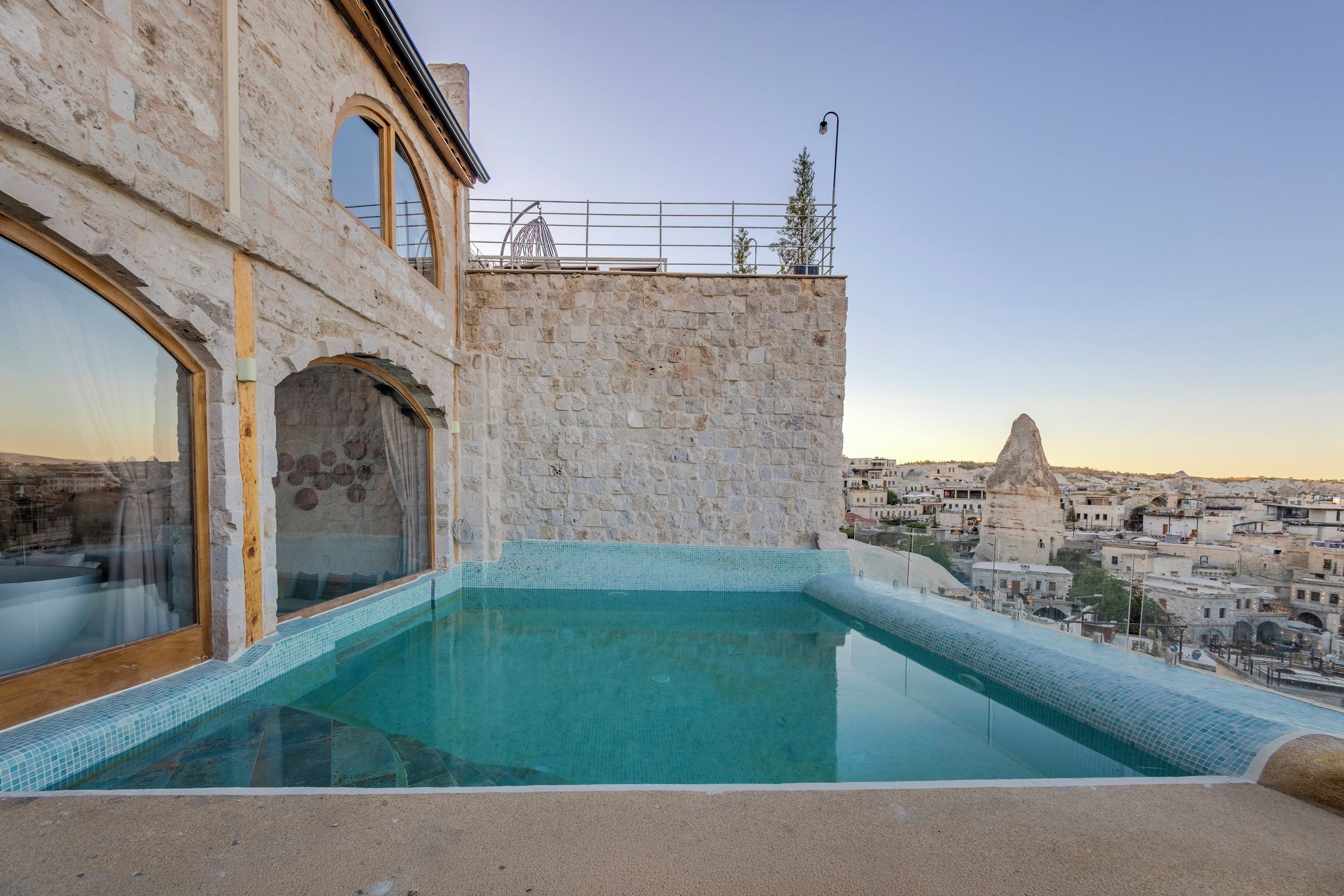 A rooftop infinity pool with a stone wall and arched windows, overlooking a cityscape with historical buildings.