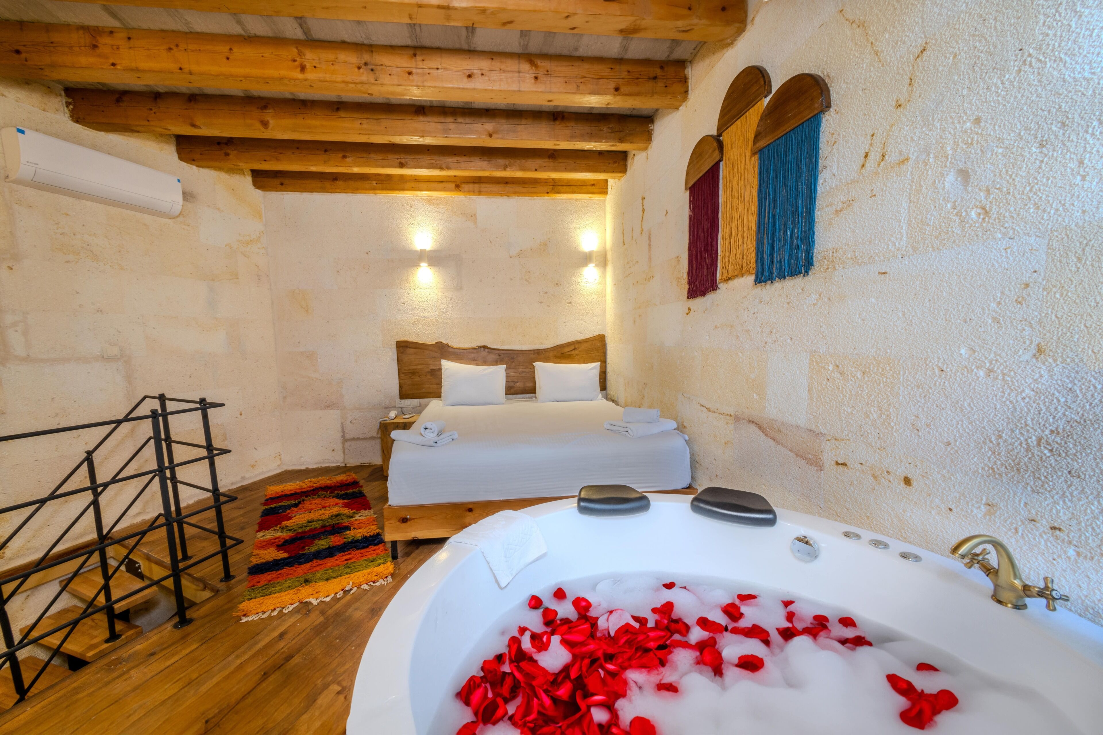A room with a wooden ceiling, a bed with white linens, a bathtub filled with red rose petals, and a colorful rug on the floor.