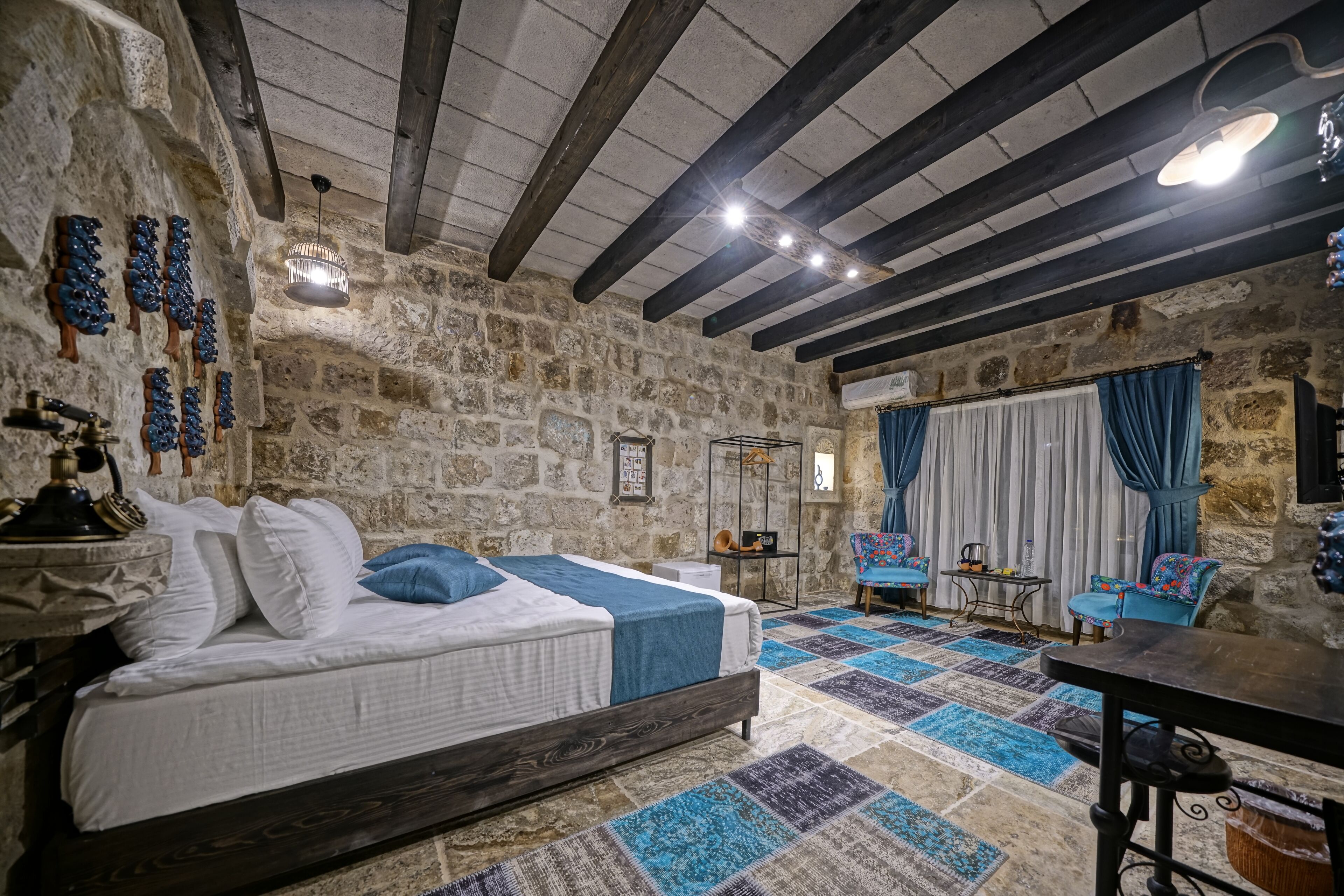 A rustic stone-walled bedroom with a wooden bed, a patterned rug, and a dining area with blue chairs.