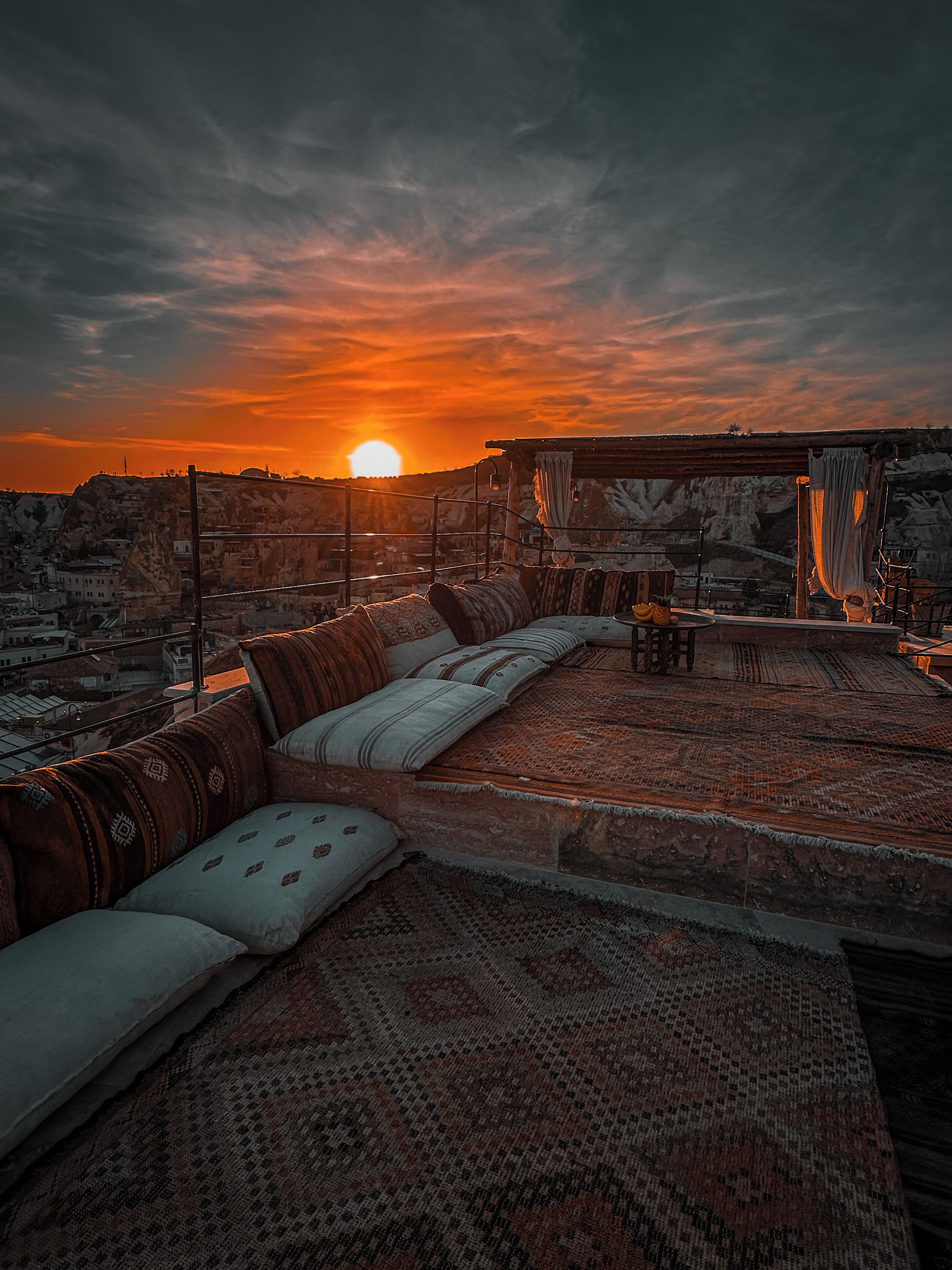 A sunset view from a rooftop terrace with a sofa and cushions, a hanging chair, and a view of the cityscape.