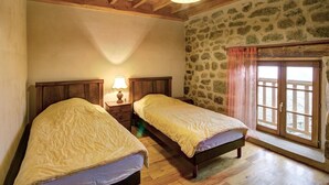 A bedroom with two single beds, wooden beams on the ceiling, a stone wall, and a window with curtains.