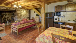 A rustic kitchen with wooden beams, a stone wall, a wooden bench, a dining table with a patterned tablecloth, and basic kitchen appliances.