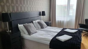A hotel room with a large bed, two bedside tables, a nightstand, a black chair, and a window with curtains. - Hotell Pausa (Klippan)