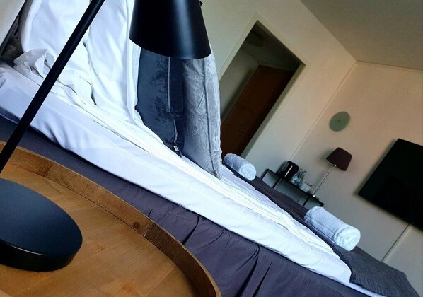 A neatly made bed with white and gray linens, a wooden headboard, and a bedside table with a lamp. A small table and a chair are visible in the background. - Hotell Pausa (Klippan)