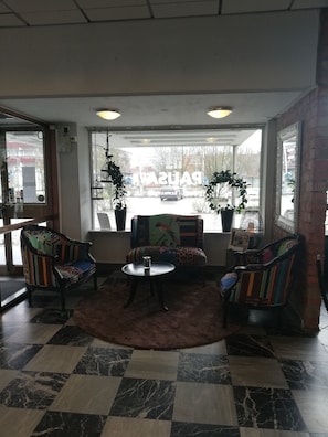 A lobby with patterned armchairs, a round table, and a view of a parking lot through large windows. - Hotell Pausa (Klippan)