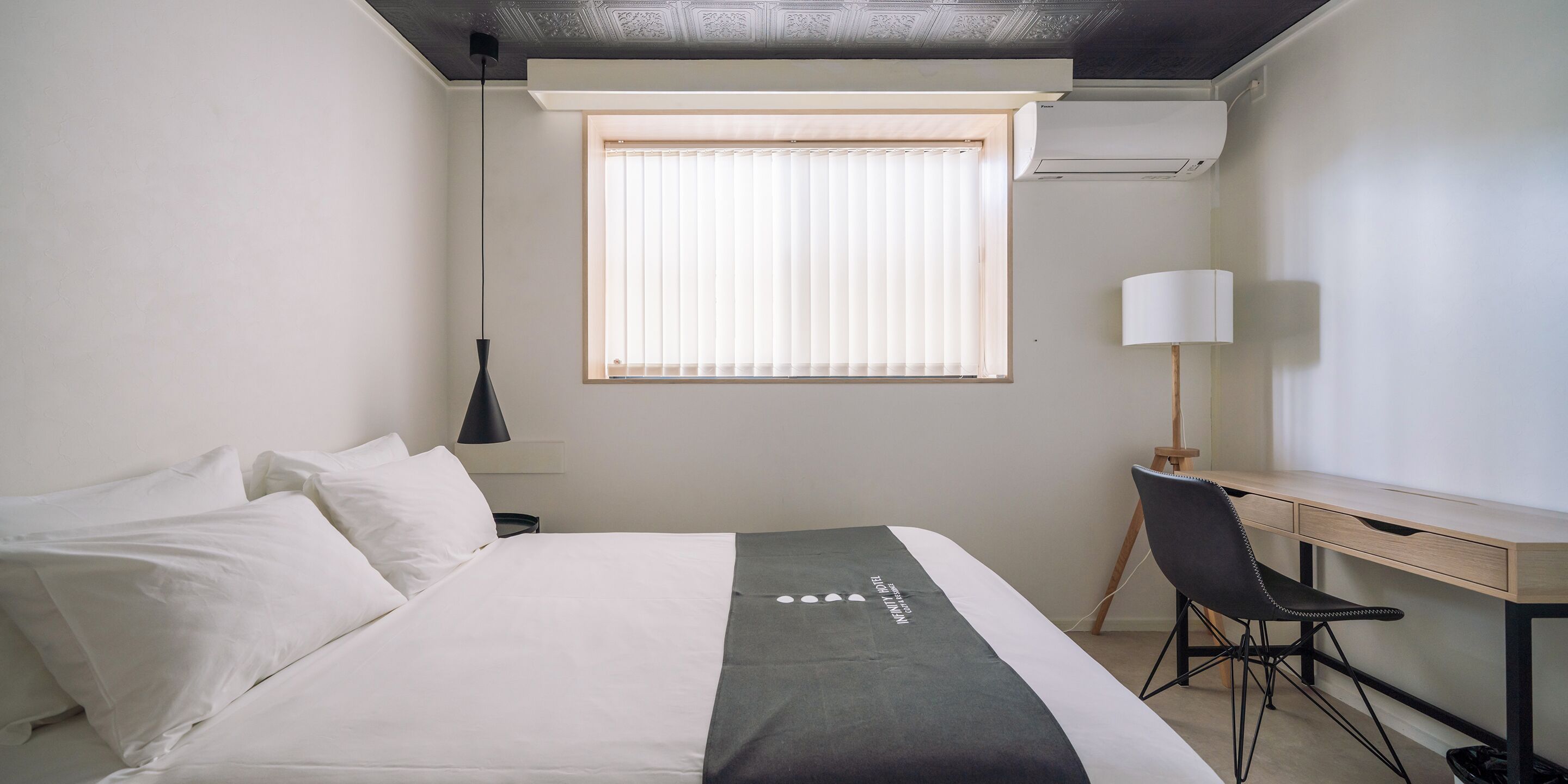 Ett hotellrum med en säng, ett skrivbord med en stol, ett fönster med persienner, en lampa och en luftkonditioneringsanläggning.