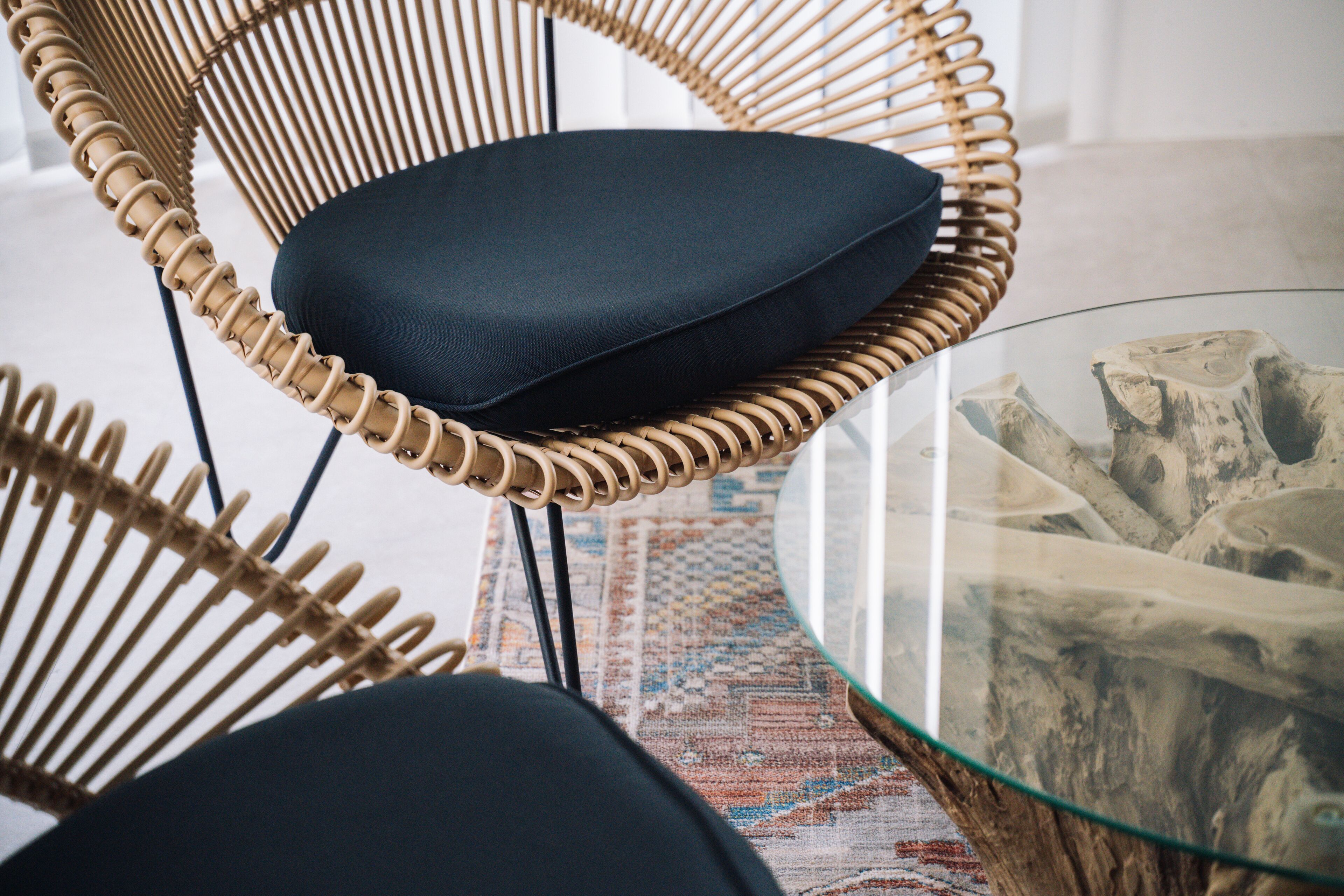 A wicker chair with a dark cushion, a glass coffee table with a wooden base, and a patterned rug on the floor.