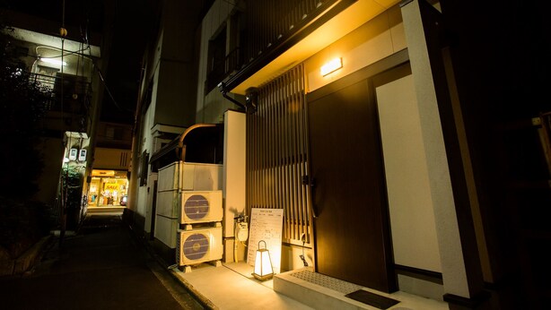 A narrow street at night with a building featuring a closed shutter door, a small illuminated sign, and two air conditioning units. - Oyado Toshi (Kyoto)