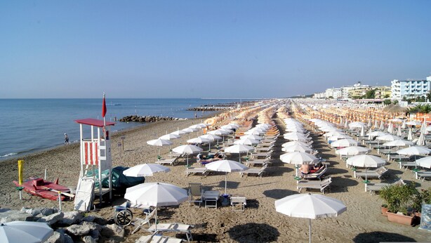 A beach with rows of white umbrellas and sun loungers, a lifeguard tower, and buildings in the background. - Holiday apartment mit Pool (Porto Santa Margherita)