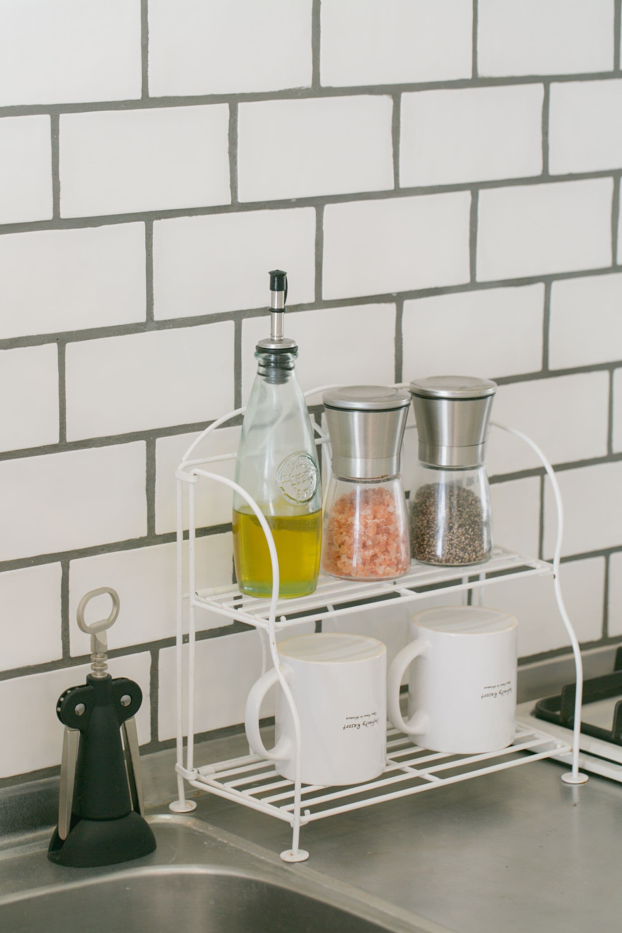 A kitchen counter with a white wire shelf holding a bottle of olive oil, two salt shakers, and two white mugs.