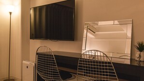 A modern bathroom with a large mirror, a flat-screen TV mounted above the mirror, and two wire chairs. - Akebono 5-C (Naha)