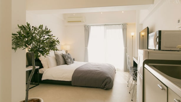 A compact bedroom with a single bed, bedside table, lamp, and a potted plant. Adjacent is a kitchenette with a microwave, sink, and cabinets. - Akebono 5-C (Naha)