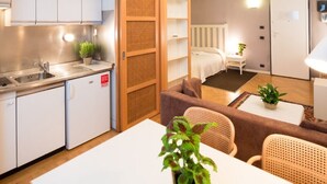 Een compacte hotelkamer met kitchenette, een bed met witte hoofdbord, een zithoekje met bank en stoelen, en een nachtkastje met een plant.