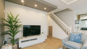 A modern living room with a flat-screen TV on a white console, a grey armchair with a patterned cushion, a staircase, and a kitchen area in the background. - Sunset Breeze Chatan  (Chatan)