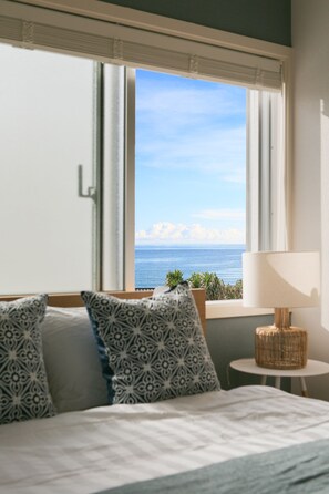 Un dormitorio con cama, almohadas estampadas, una mesita de noche con lámpara y vista al océano a través de una ventana.