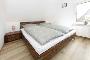 A wooden bed with white bedding in a room with a white radiator and a window with a plant on the sill.