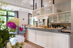 Una cocina moderna con armarios blancos, estantes de vidrio que exhiben botellas y copas de vino, y vistas del jardín exterior a través de grandes ventanales.