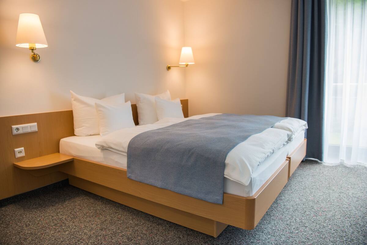 A hotel room with a bed, two bedside lamps, a nightstand, and a window with curtains.