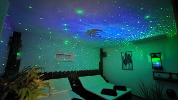 Habitación con techo que presenta una proyección de luces tipo cielo estrellado, cama con ropa de cama blanca y televisor montado en la pared.