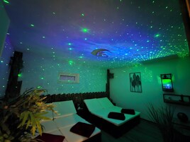 Quarto com teto exibindo uma decoração de estrelas, cama com roupa de cama branca e televisão fixada na parede.
