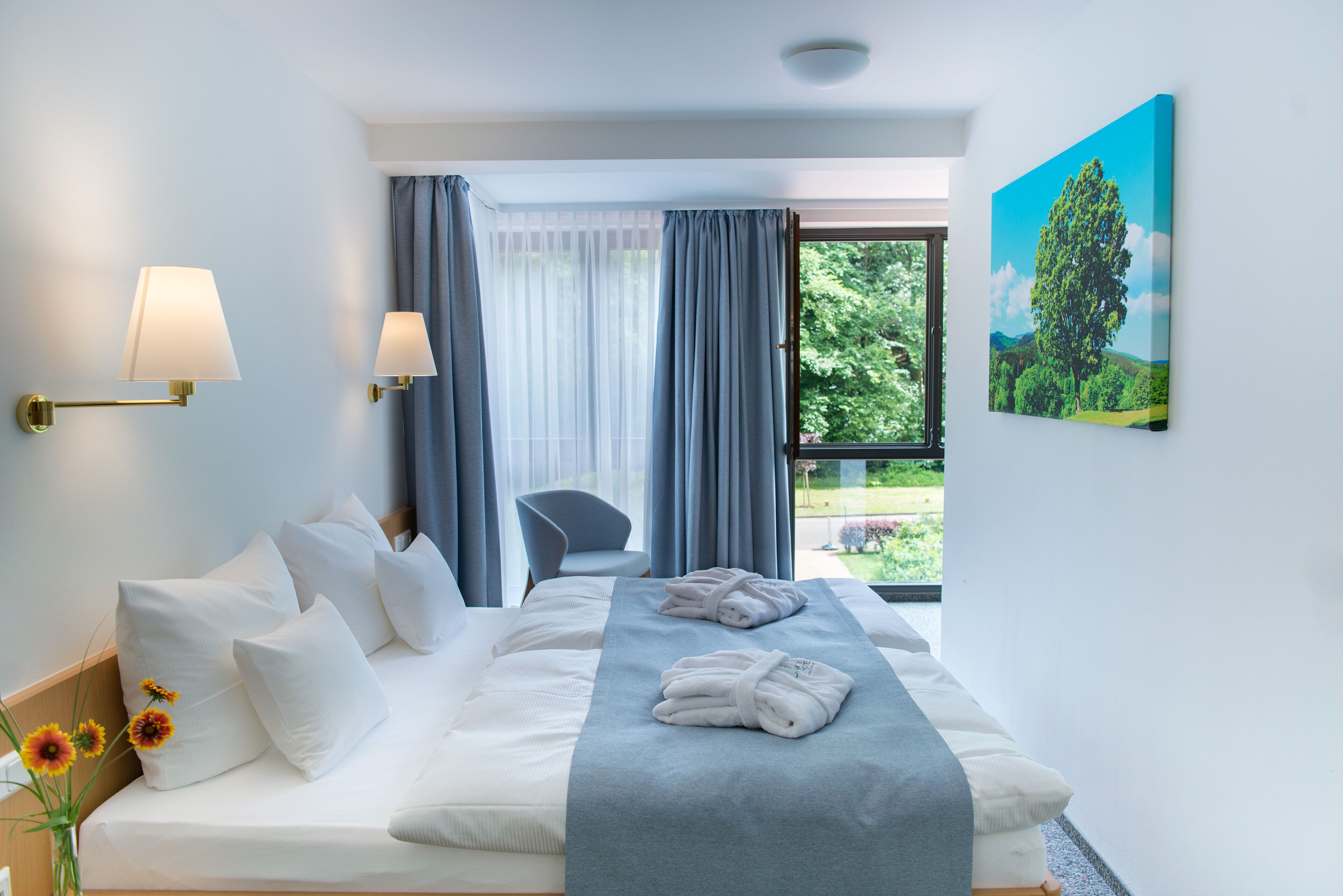 Habitación de hotel con cama, almohadas, lámpara de noche, una silla, ventana con cortinas y un cuadro en la pared.
