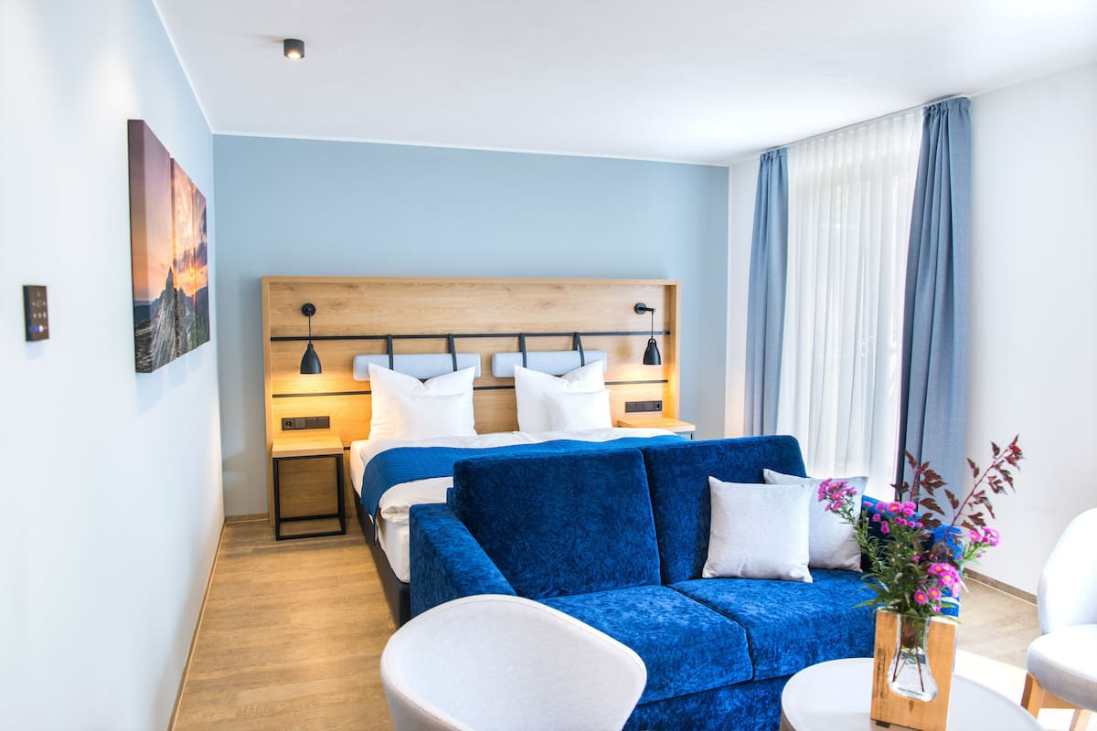 A modern hotel room with a wooden headboard bed, a blue sofa, a white armchair, and a small table with a vase of flowers.