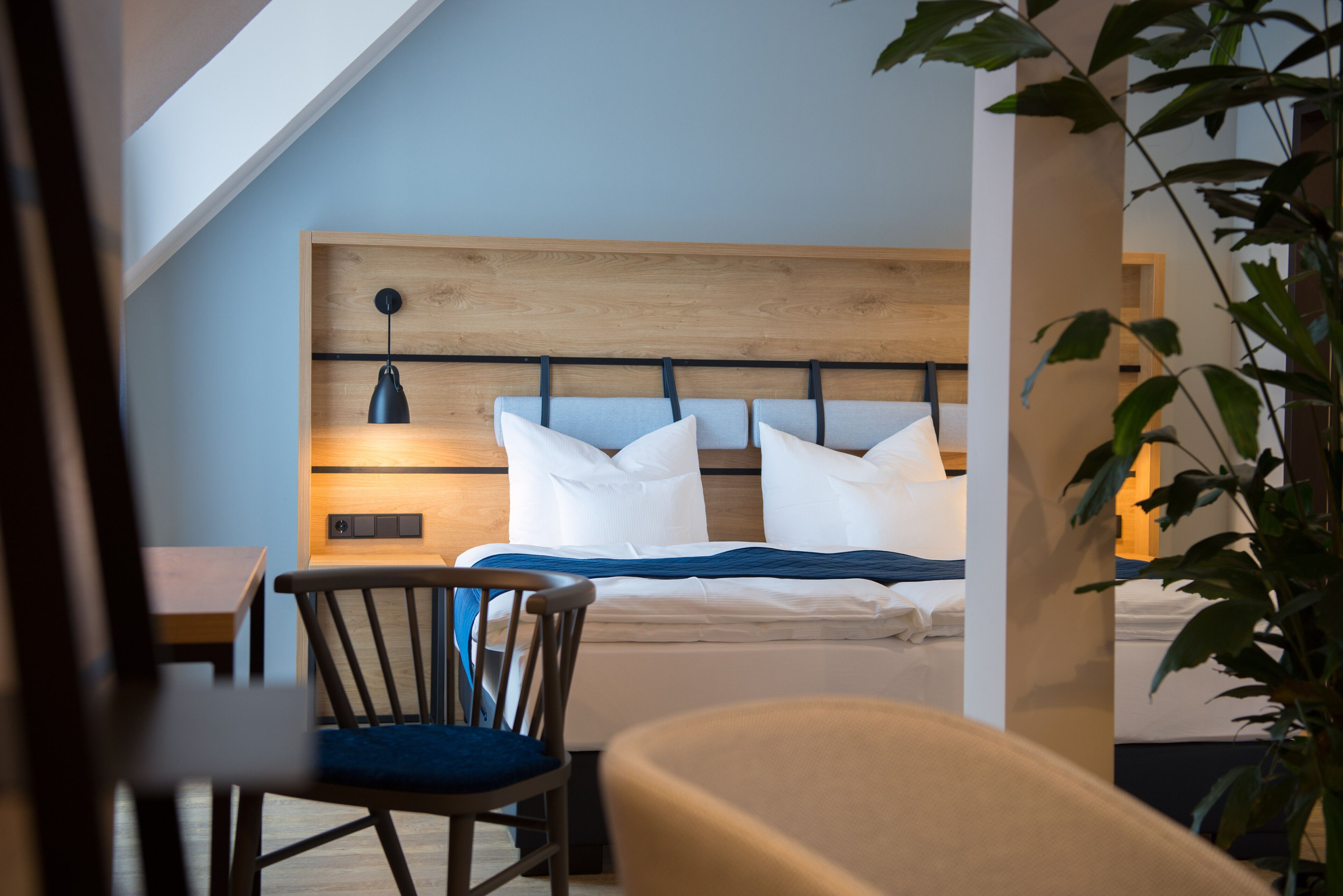 A bedroom with a sloped ceiling, a wooden headboard, a bed with white bedding, a desk with a chair, and a potted plant.
