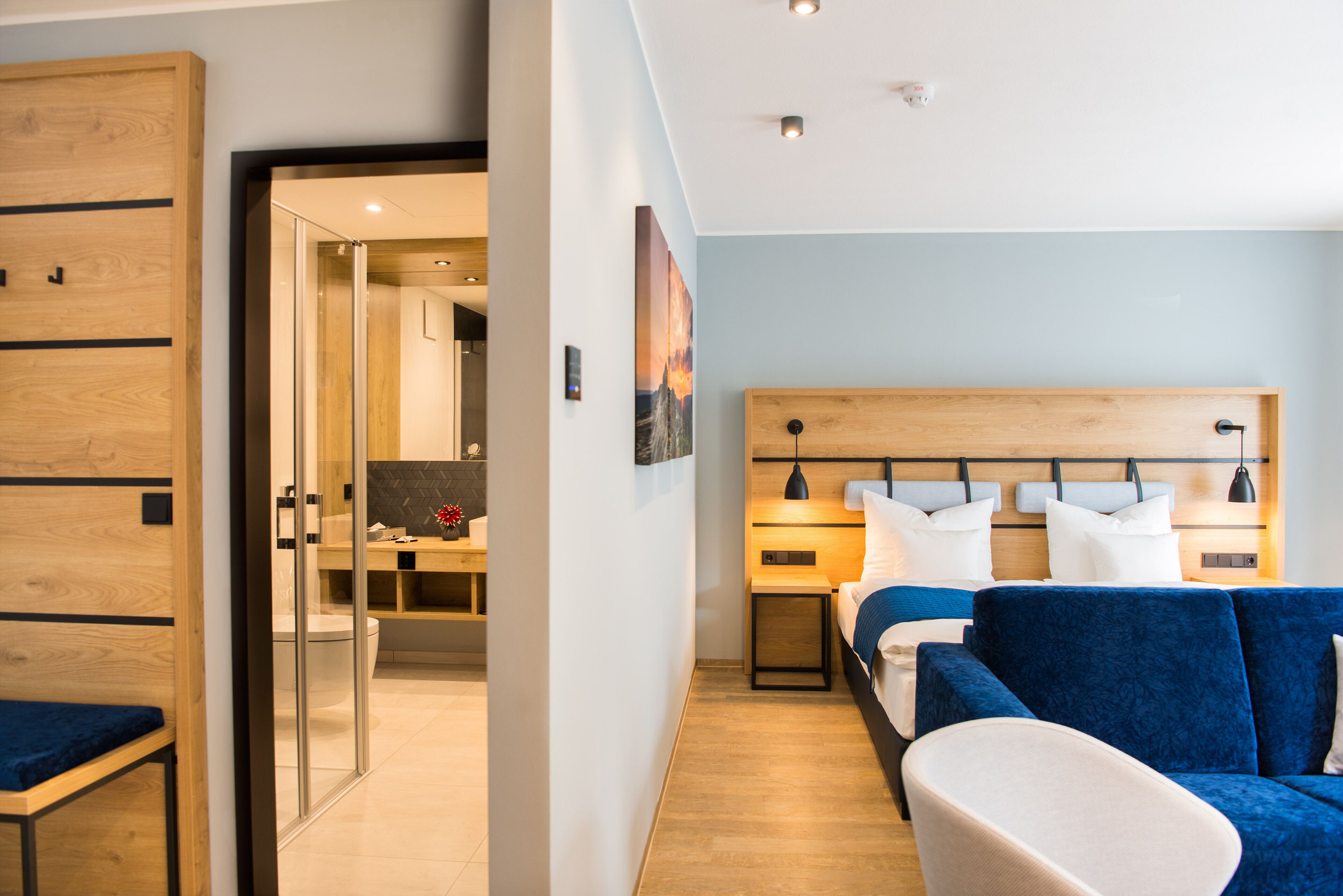 A modern hotel room with a wooden headboard, a bed with blue bedding, a white armchair, and a desk with a computer.