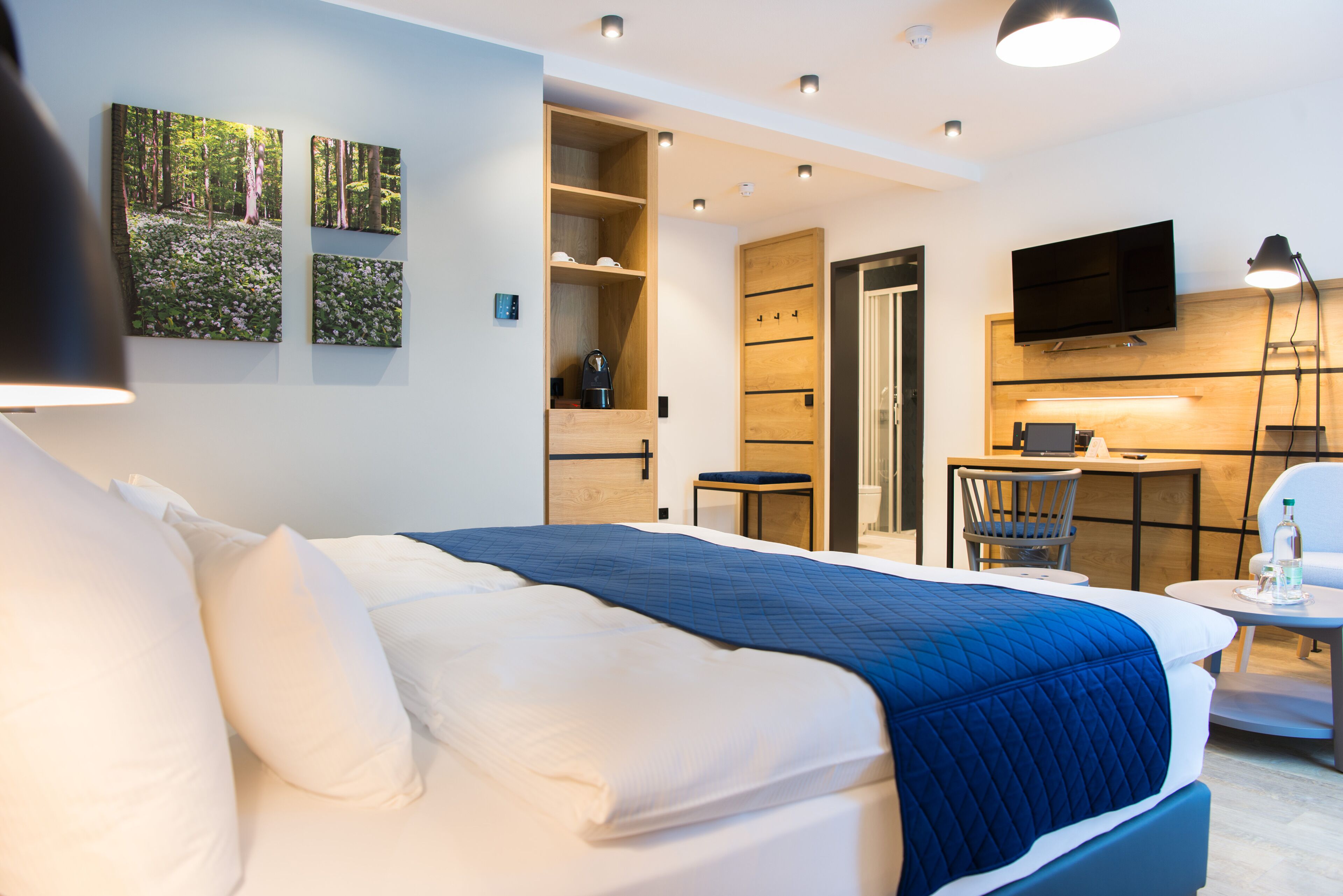 A modern hotel room with a large bed, a desk, a chair, and a television. There are wall-mounted shelves and a coffee machine.