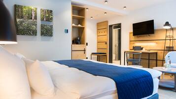 A modern hotel room with a large bed, a desk, a chair, and a television. There are wall-mounted shelves and a coffee machine.