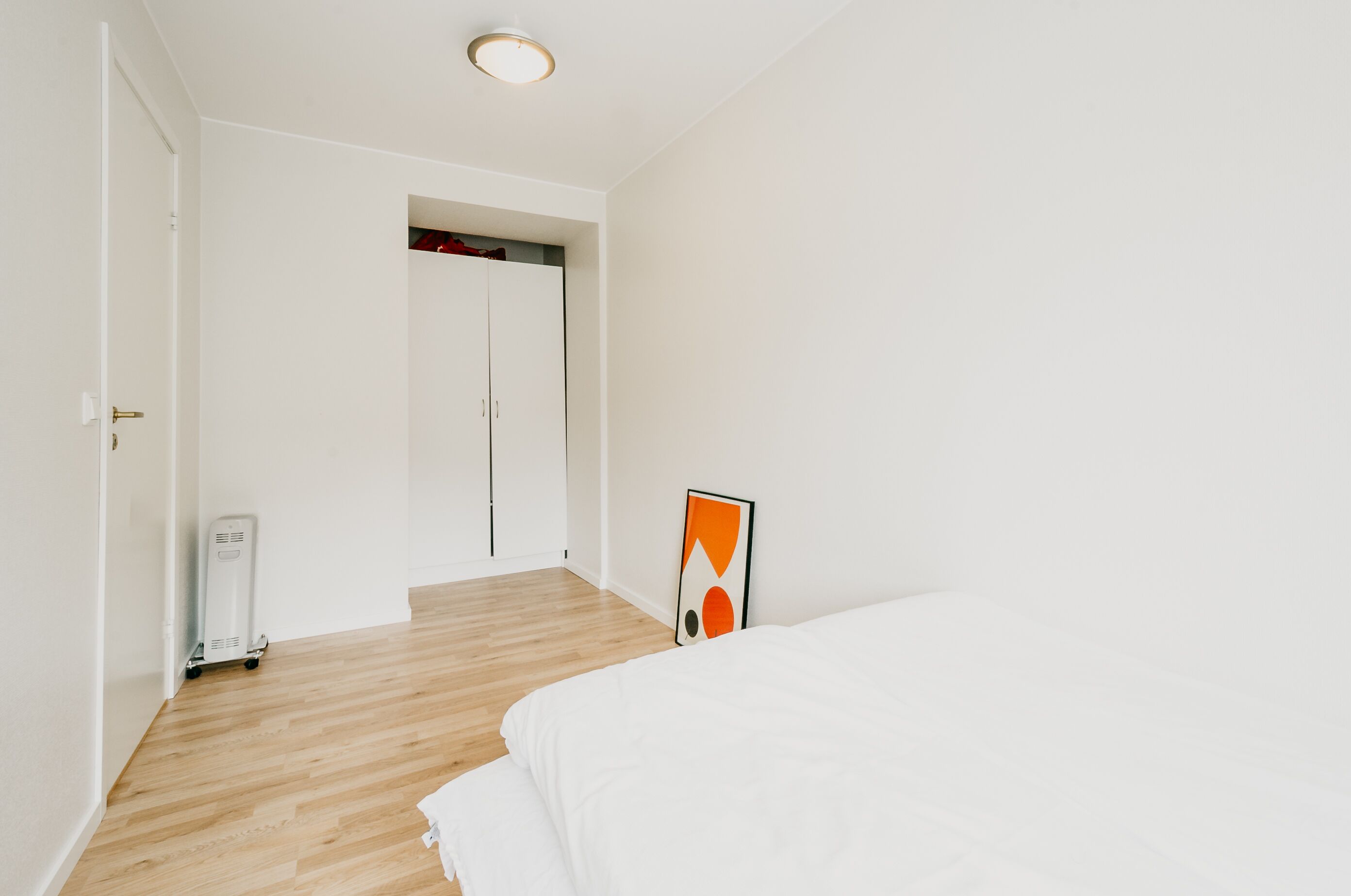 Een slaapkamer met een houten vloer, een wit bed, een witte kledingkast en een ingelijste foto aan de muur.