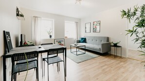 Ett modernt vardagsrum med en matplats, inrett med en grå soffa, svarta stolar, ett litet soffbord och ett matbord med stolar. Det finns två fönster med gardiner, en taklampa och en vägghylla med växter.